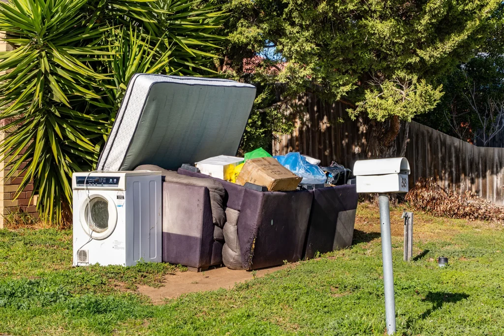 Hard rubbish left on the front yard garden of a suburban property featuring an old washing machine, broken furniture, mattresses, and discarded household items.